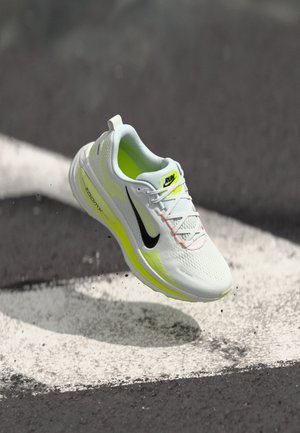 White athletic shoe with neon yellow accents, mesh upper, and black logo. Features a cushioned sole and reflective detailing, suspended above a surface.