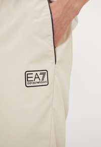 Beige lättviktstyg med en slät yta, med en svart dragkedja och en logotyp-patch märkta "EA7 Emporio Armani" på vänster sida.