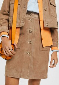 Suède knoopsluiting tan rok gecombineerd met een bijpassende tan jas en een gelaagd oranje shirt, met een kleine oranje schoudertas.