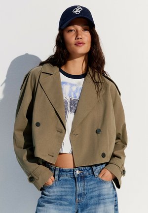 Ung kvinde iført en kort olivengrøn jakke, grafisk crop top, blå jeans og en navyblå baseballkasket poserer med hænderne i lommerne.