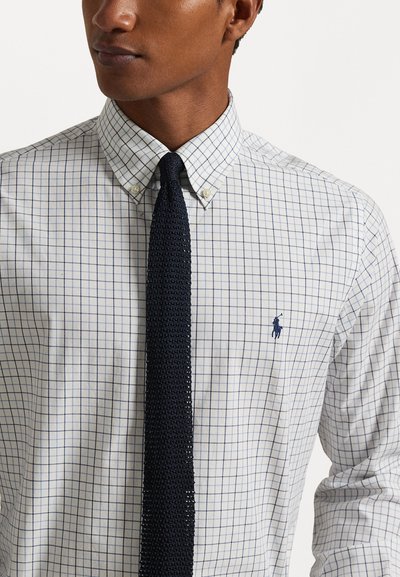 Camisa blanca de botones con cuadros azules y un pequeño logo bordado. Una corbata de punto en un oscuro azul marino añade contraste al conjunto.