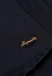 Sort ribbet stof med en glat overflade, der har et guld broderet "Superdry" logo på den ene side.