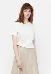 Jonge vrouw met rood haar, gekleed in een witte shirt met korte mouwen en een beige rok, staand tegen een effen witte achtergrond.