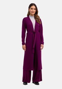 Long cardigan violet avec une ceinture à nouer, présentant des manches longues et une coupe décontractée, porté par-dessus un haut gris et un pantalon large.