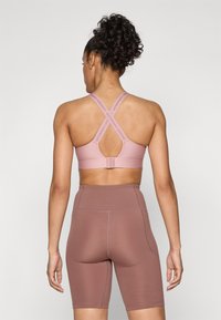 Rosa treningsbh med en nøkkelhull-design bak, justerbare stropper og logo-detaljer, kombinert med brune høytaljede shorts. Glatt tekstur.