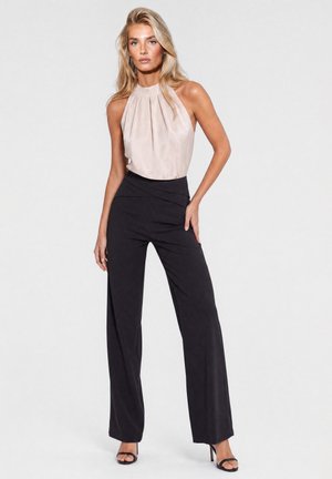 Woman standing in beige sleeveless top and high-waisted black wide-leg pants with black heeled sandals against a plain background.