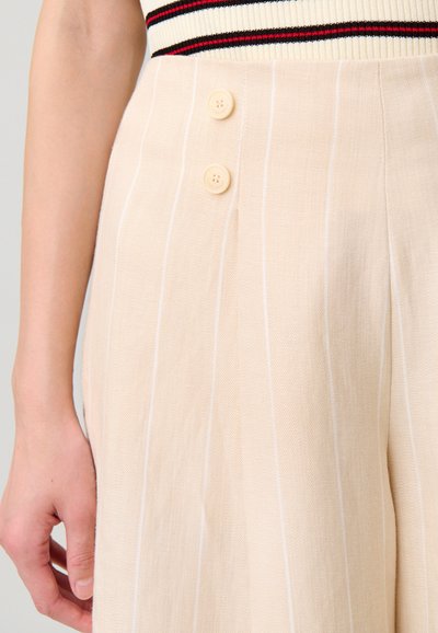 Pantaloni beige plissettati con strisce verticali bianche. Presentano due bottoni a contrasto in vita, realizzati in un tessuto leggero.