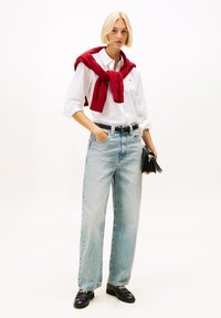 White button-up shirt, red knitted sweater tied around the shoulders, light blue wide-leg jeans, and black loafers, holding a small black bag.