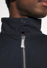 Veste bleu marine avec un col montant, dotée de deux boutons noirs et d'une fermeture éclair argentée avec un porte-étiquette. La texture du tissu est lisse.