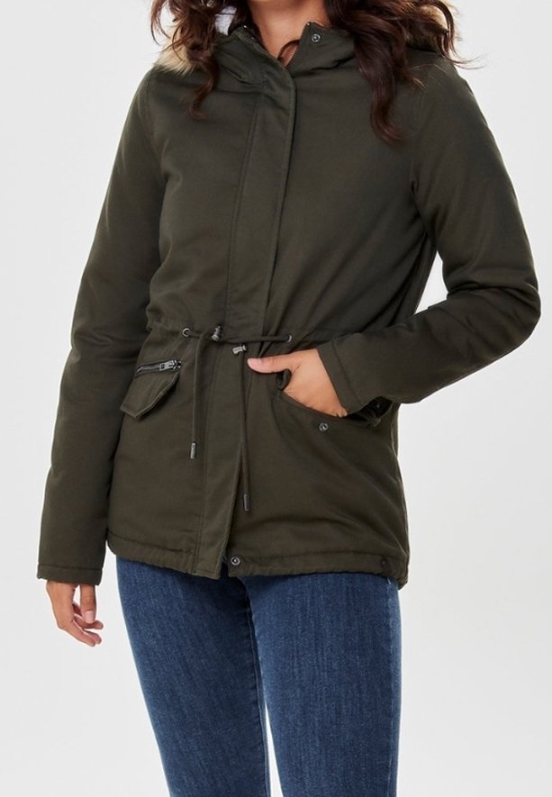 Femme portant une veste d'hiver à capuche vert olive avec taille ajustable par cordon et un jean bleu, main dans la poche, sur un fond uni.