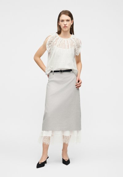 Femme portant un chemisier blanc à manches bouffantes en œillets, une jupe midi gris clair avec ourlet en dentelle, une ceinture noire et des chaussures à talons pointues noires, debout devant un fond blanc.