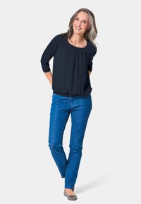 Schwarze Bluse mit dreiviertel Ärmeln, plissierter Vorderseite, kombiniert mit blauen Jeans und grauen Ballerinas. Frau steht mit Händen auf den Hüften und lächelt.