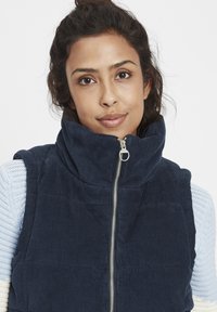 Mörkblå kordflortsväst med hög krage, dragkedjestängning och fickor med lappar. Bärs över en ljusblå texturerad tröja.