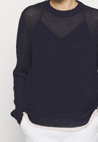 Nahaufnahme einer Person, die einen strukturierten dunkelblauen Strickpullover über einem schwarzen Top trägt, kombiniert mit weißen Hosen.