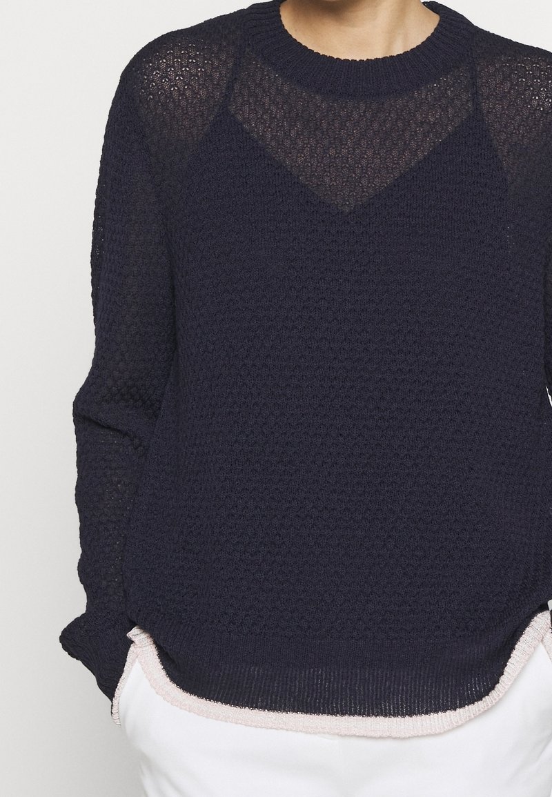 Nahaufnahme einer Person, die einen strukturierten dunkelblauen Strickpullover über einem schwarzen Top trägt, kombiniert mit weißen Hosen.