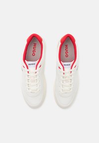 White sneakers with red accents, featuring a smooth textile upper, round toe, and decorative stitching. Shoe tongue and insole labeled "HUGO."