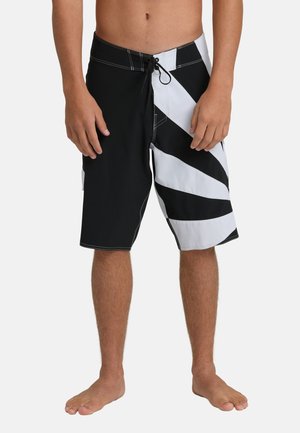 Man draagt zwart-witte boardshorts met een diagonaal strepenpatroon en staat blootsvoets tegen een effen achtergrond.
