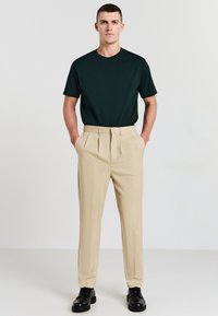 Un uomo sta in piedi di fronte con le mani nelle tasche di pantaloni beige plissettati, indossando una maglietta nera e scarpe eleganti nere su uno sfondo semplice.