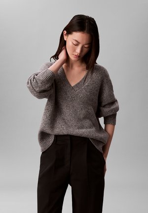Junge Frau in lockerem grauem V-Ausschnitt-Pullover und schwarzen Hosen, mit einer Hand am Nacken und gesenktem Kopf vor einfarbigem grauem Hintergrund.