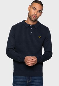 Námořnická modrá pletená polo svetr s strukturovaným vzorem, tři knoflíky u límce a malé žluté logo na hrudi. Měkká tkanina a dlouhé rukávy.