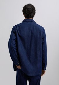 Man met kort krullend haar, gekleed in een donkerblauwe spijkerjack en bijpassende spijkerbroek, staand met zijn rug naar de camera tegen een effen lichte achtergrond.