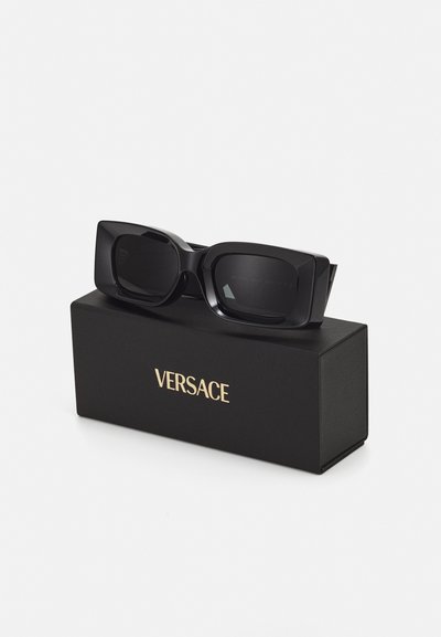 Lunettes de soleil noires rectangulaires avec des montures épaisses, présentant des verres teintés, posées sur une boîte noire avec le logo "Versace" en or.