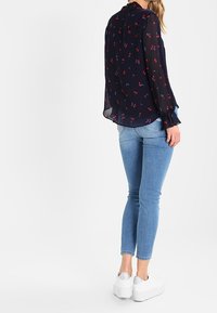 Donna che indossa una blusa floreale scura e trasparente, jeans skinny azzurri chiari e sneakers bianche con la piattaforma, in piedi con le spalle rivolte.