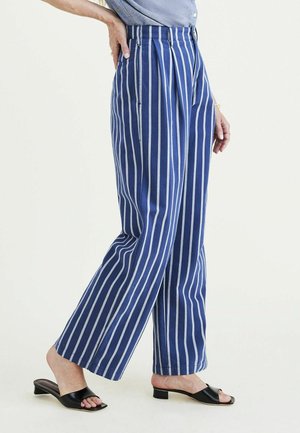 Femme portant un pantalon large rayé verticalement bleu et blanc et des sandales noires à bout ouvert et talon bas, debout avec une main sur la hanche.