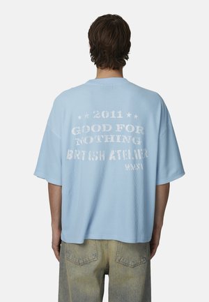Maglietta oversize azzurro chiaro con texture a coste. Presenta un testo stampato bianco sul retro, tra cui "GOOD FOR NOTHING" e "BRITISH ATELIER".