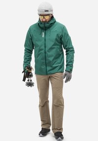 Veste étanche verte avec capuche, pantalon beige, gants gris et bottes noires. Tenant des bâtons de trekking, portant un bonnet gris et des lunettes de soleil.