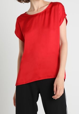 Kobieta ubrana w jaskrawoczerwoną bluzkę z krótkim rękawem i okrągłym dekoltem oraz czarne spodnie z białymi bocznymi paskami na jednolitym tle.