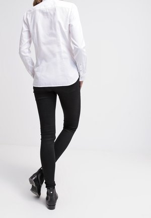 Person iført en hvid skjorte med lange ærmer, sorte skinny bukser og sorte ankelstøvler, der står med det ene ben krydset bag det andet.
