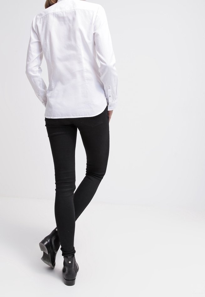 Personne portant une chemise blanche à manches longues, un pantalon skinny noir et des bottines noires, debout avec une jambe croisée derrière l'autre.