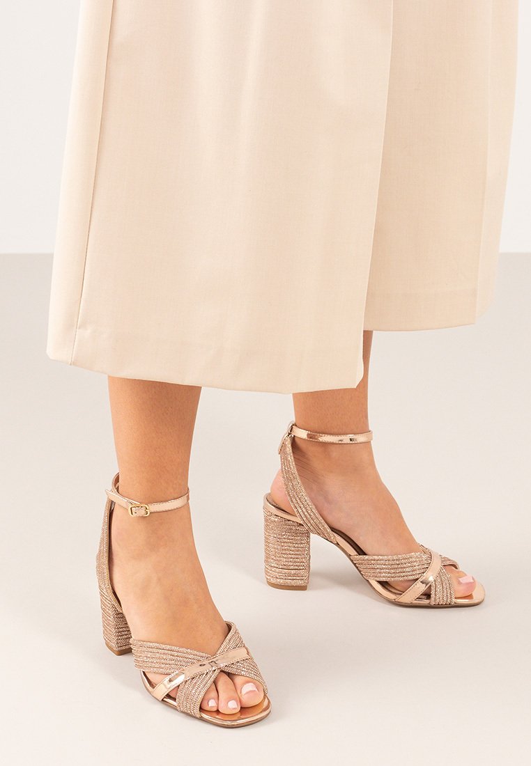 Sandalias de tacón alto beige con una tira delantera anudada, tira de tobillo trenzada texturizada y un bloque de tacón robusto. Plantilla suave y diseño de punta abierta.