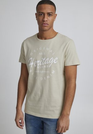 Beige bomulds t-shirt med korte ærmer, rund halsudskæring, med en hvid trykt grafik, der står "Heritage Blend" i midten.