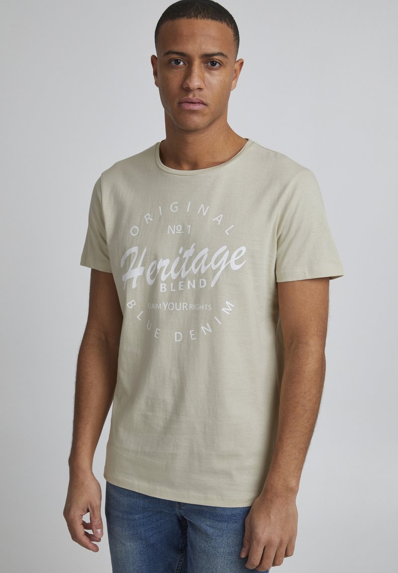 Beige bomull t-skjorte med korte ermer og rund hals, med et hvitt trykt grafikk som leser "Heritage Blend" i midten.