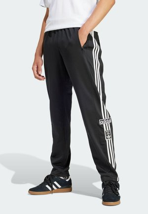 Persoon draagt zwarte Adidas trainingsbroek met witte strepen en logo, gecombineerd met zwarte Adidas sneakers en wit shirt, staat met hand in zak.