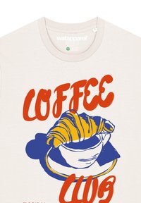 Crème-kleurig t-shirt met een grafisch ontwerp van een croissant boven een koffiekop met de tekst "COFFEE CLUB" in het rood. Eenvoudig, gedurfd ontwerp.