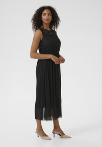 Black sleeveless dress featuring a lace bodice, flowing skirt, and sheer bottom. Paired with beige slingback heels.