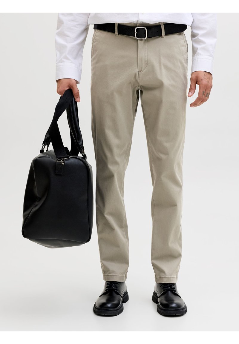 Άνδρας που φοράει μπεζ παντελόνι, μαύρη ζώνη, μαύρα παπούτσια και λευκό πουκάμισο, κρατώντας μια μαύρη τσάντα duffel στο αριστερό του χέρι μπροστά σε λευκό φόντο.