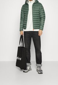Grønne pufferjakke med hætte, lynlåslukning og orange detaljer. Hvid langærmet skjorte, sorte jeans, grå sneakers og sort totebag.