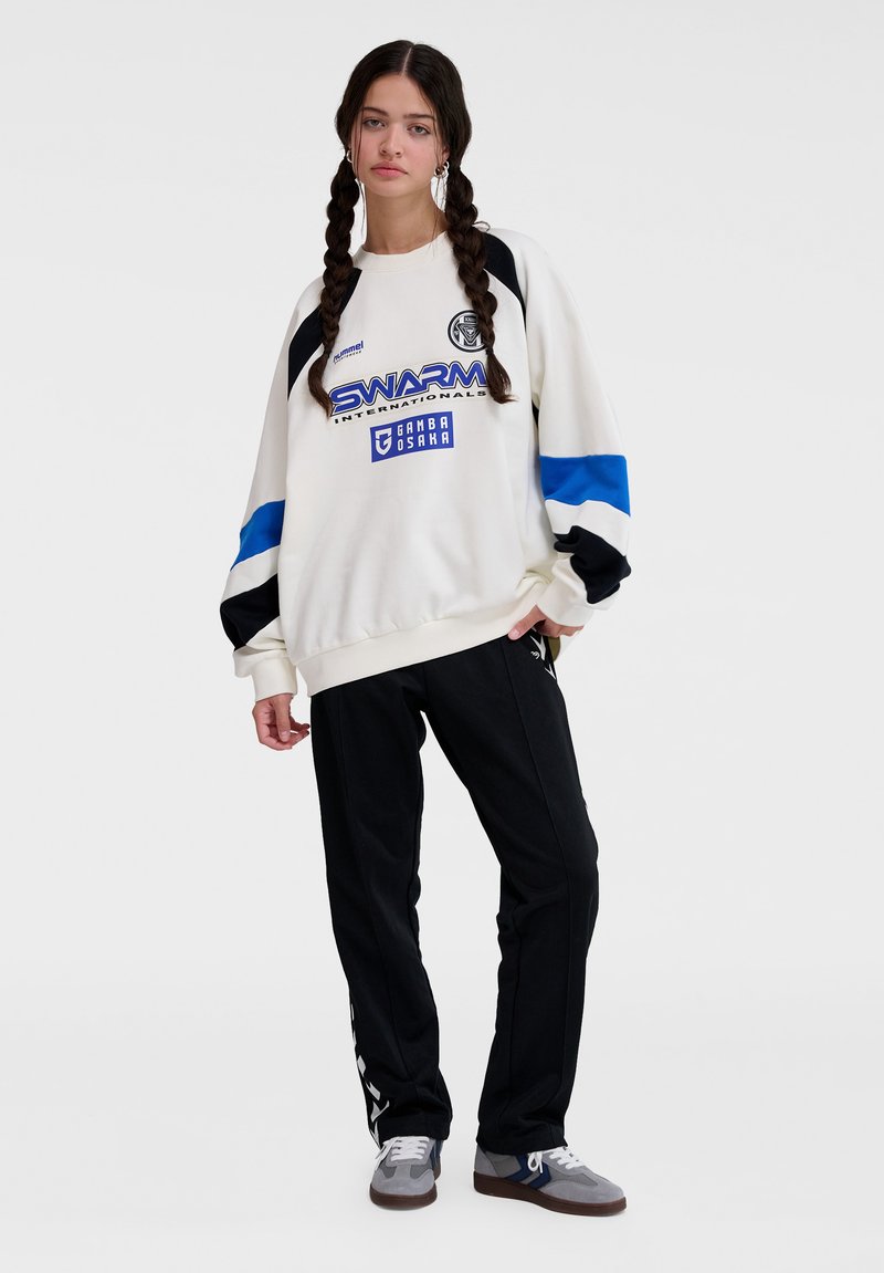 Överdimensionerad krämfärgad sweatshirt med en blå och svart design, med logotyp framtill; matchad med svarta byxor och grå sneakers.