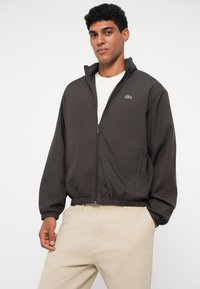 Giacca zip-up marrone scuro con polsini e orlo elasticizzati, caratterizzata da un piccolo logo verde. Indossata su una camicia chiara e pantaloni beige.