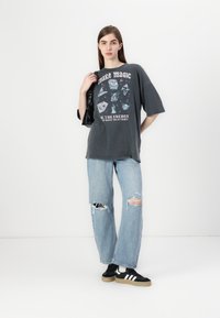 T-shirt graphique noire avec le texte "Make Magic" et des motifs, assortie à un jean déchiré bleu clair et des baskets noires avec des rayures blanches.