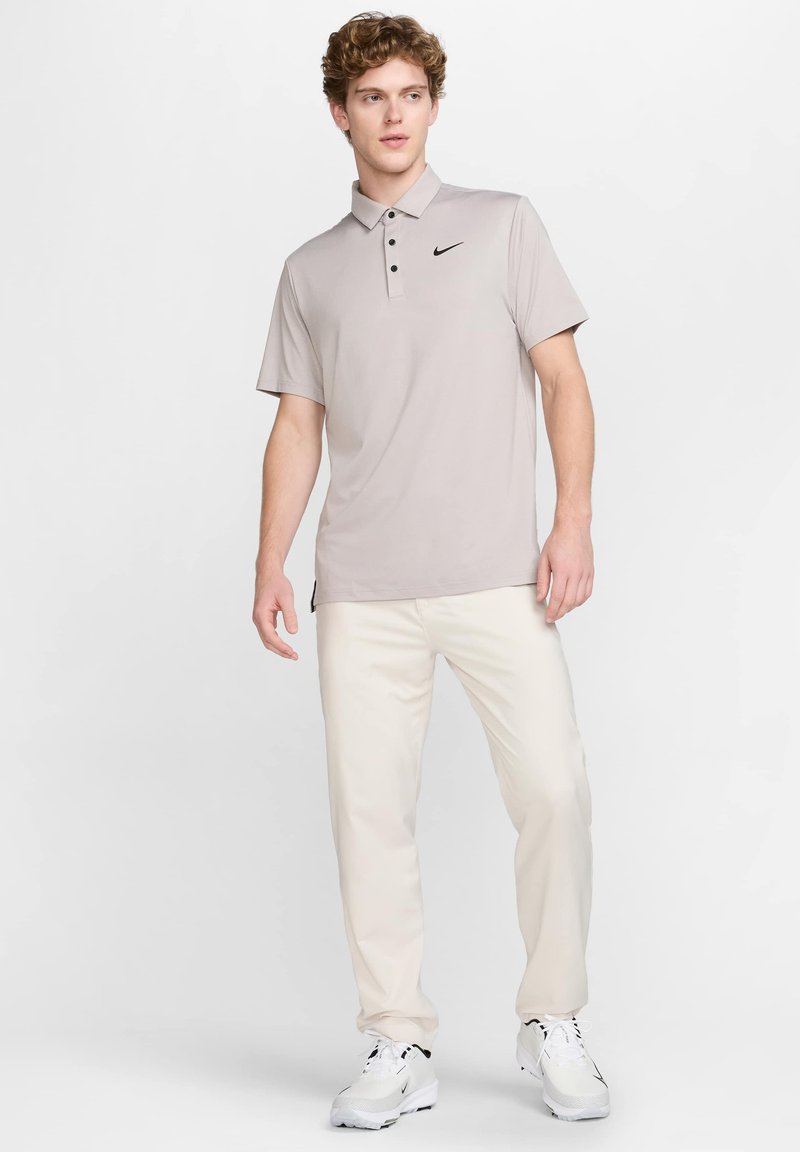 Lys grå polo t-shirt med en subtil glans, tre-knaps placket og et lille sort logo. Kombineret med kridtfarvede bukser og hvide sneakers.