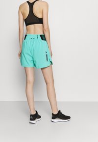 Persona vestida con un sujetador deportivo negro, pantalones cortos turquesa de Adidas Terrex y zapatillas negras de Adidas, de espaldas frente a un fondo blanco.