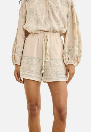 Top de manga larga beige bordado y pantalones cortos a juego con cintura de cordón, usados por una persona de pie sobre un fondo liso.