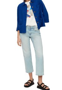 Blaues Leinenjackett mit großen Knöpfen über einem weißen Grafik-T-Shirt, kombiniert mit hellblauen cropped Jeans und schwarzen Sandalen mit Riemen.