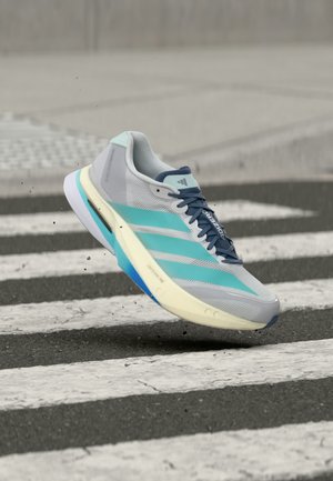 Zapatilla de correr gris claro y turquesa con cordones oscuros en el aire sobre un paso de peatones en blanco y negro sobre asfalto.