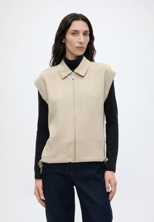 Frau mit langen dunklen Haaren, die eine beige ärmellose Reißverschlussweste über einem schwarzen Langarmshirt und dunklen Jeans trägt, vor einem schlichten Hintergrund stehend.